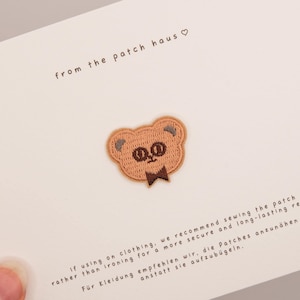 Può includere: Una toppa ricamata a forma di testa di orso nei toni del marrone e beige con un piccolo papillon, centrata su un cartoncino bianco. Il cartoncino riporta la scritta from the patch haus e istruzioni che consigliano di cucire la toppa.