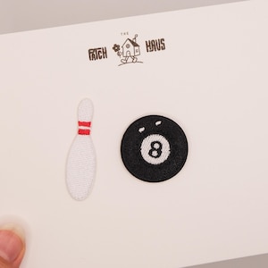 Könnte beinhalten: Zwei gestickte Aufnäher auf einer weißen Karte mit dem Text THE PATCH HAUS und einem kleinen Haus-Symbol. Ein Aufnäher ist eine weiße Bowlingkugel-Kegel mit zwei roten Streifen, der andere ist eine runde schwarze Acht-Ball-Kugel mit einer weißen Acht.