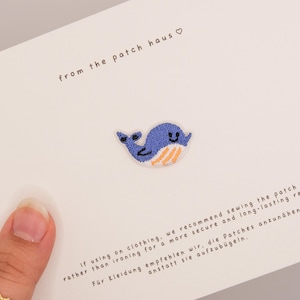Könnte beinhalten: Ein kleiner gestickter Wal-Aufnäher mit blauem Körper und orangefarbenen Streifen am Bauch, präsentiert auf einer weißen Karte mit dem Text from the patch haus und Anweisungen zum Annähen des Aufnähers auf Kleidung.