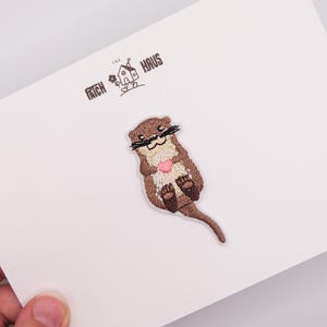 Op de afbeelding: Een geborduurde patch met een bruine otter die een klein roze hartje vasthoudt, geplaatst op een witte kaart met de tekst THE PATCH HAUS en een klein huis-icoon in het zwart gedrukt boven het otterontwerp.