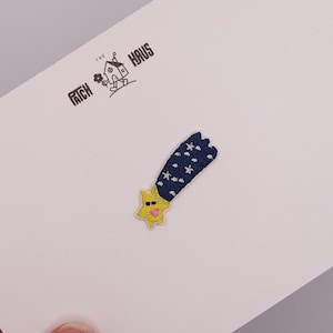 Puede incluir: Un parche bordado que muestra una estrella amarilla con nariz rosa y ojos negros, dejando una estela de cometa azul oscuro decorada con pequeñas estrellas blancas. El texto THE PATCH HAUS está impreso en negro sobre una tarjeta blanca.