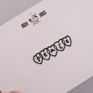 Puede incluir: Un parche bordado con cinco segmentos blancos en forma de corazón, cada uno con una letra negra que forma la palabra PEACE. Sobre el parche, el texto THE PATCH HAUS está impreso en negro con un pequeño icono de una casa sosteniendo una flor.