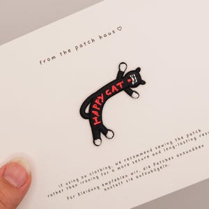 May include: An embroidered black cat patch with white paws and the red text HAPPY CAT curved along its body. The patch is attached to a white card featuring the text from the patch haus and instructions for sewing the patch onto clothing.