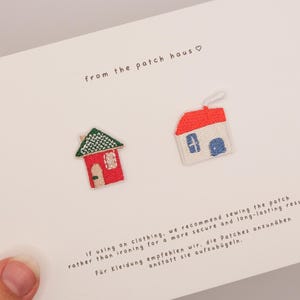 May include: Two embroidered house patches on a white card with the text from the patch haus. One patch is a red house with a green roof and a tan door. The other is a white house with a red roof, blue windows, and a white chimney with smoke.