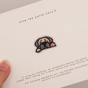Puede incluir: Un parche bordado que muestra un perro de color beige y negro con una pequeña flor roja, centrado en una tarjeta blanca con el texto from the patch haus e instrucciones que recomiendan coser el parche en lugar de plancharlo.