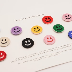 Op de afbeelding: Een verzameling van elf ronde geborduurde patches met smiley-gezichten in verschillende kleuren, waaronder rood, blauw, geel, groen, paars, roze, zwart en crème. De tekst from the patch haus staat gedrukt op de witte kaart.