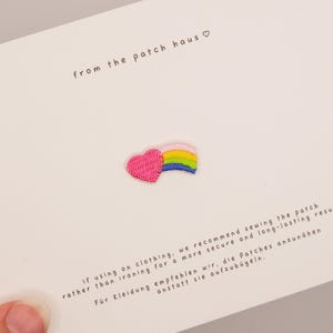 Puede incluir: Un pequeño parche bordado con un corazón rosa brillante y un arcoíris multicolor. El parche está sujeto a una tarjeta blanca con el texto from the patch haus e instrucciones para coser el parche en la ropa en inglés y en alemán.