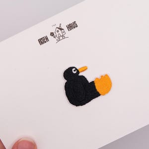 May include: An embroidered patch featuring a black bird with a bright orange beak and webbed foot. The patch is attached to a white card printed with the text THE PATCH HAUS and a small line drawing of a house with legs holding a flower.