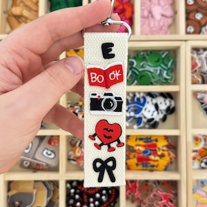 May include: A cream-colored fabric keychain strap featuring five embroidered patches stacked vertically: a black letter E, a red open book with the text "BOOK", a black camera, a red heart with a face and limbs, and a black bow.