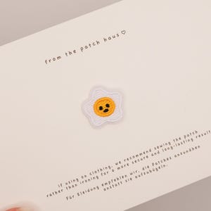 Könnte beinhalten: Ein gestickter Aufbügler in Form eines Spiegeleis mit weißem Rand und gelber Mitte, die ein Gesicht mit schwarzen Punkten zeigt. Der Aufnäher ist auf einer weißen Karte befestigt, die mit dem Text from the patch haus und Anleitungen bedruckt ist.