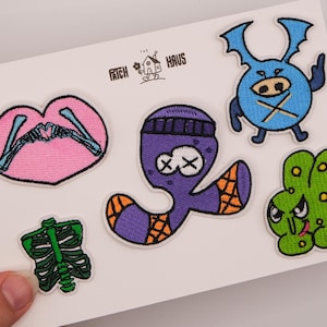 Peut inclure: Un ensemble de cinq écussons brodés sur une carte blanche avec le texte THE PATCH HAUS. Les motifs incluent un cœur rose avec des mains squelettiques, une cage thoracique verte, une créature violette avec un bonnet, un personnage bleu ailé et un monstre vert.