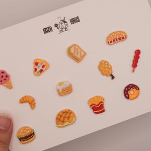 Może przedstawiać: Zestaw dwunastu małych haftowanych naszywek z motywem jedzenia na białej karcie z napisem The Patch Haus. Wzory obejmują rożek lodowy, kawałek pizzy, kanapkę, naleśniki, rogalik, burgera, pączka, frytki oraz inne przekąski.