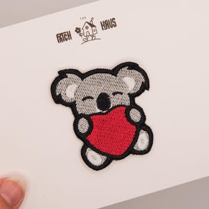 May include: An embroidered patch featuring a gray koala with a black outline holding a bright red heart. The patch is attached to a white card with the text THE PATCH HAUS and a small illustration of a house with a flower printed at the top.