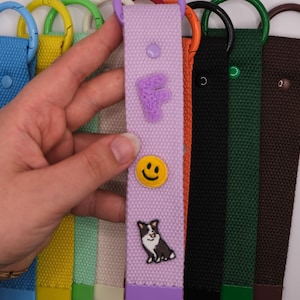 Könnte beinhalten: Eine Sammlung von Stoff-Schlüsselanhängern in verschiedenen Farben, darunter Blau, Gelb, Hellgrün, Beige, Lavendel, Orange, Schwarz, Dunkelgrün und Braun. Jedes Band verfügt über einen passenden D-Ring aus Metall und einen kleinen Zieraufnäher.