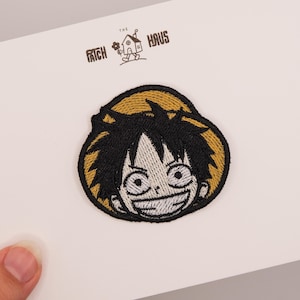 May include: An embroidered patch featuring the face of a cartoon character with black hair and a wide grin, wearing a yellow straw hat. The patch is displayed on a white card with the text THE PATCH HAUS and a small house icon printed at the top.