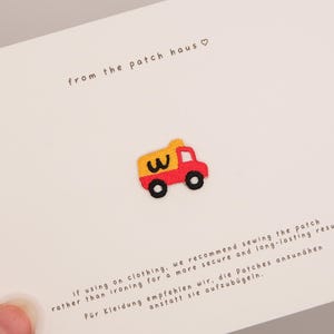 Könnte beinhalten: Ein kleiner gestickter Aufnäher in Form eines roten und gelben Kipplasters mit schwarzen Rädern und einem schwarzen Buchstaben W an der Seite. Der Aufnäher ist auf einer weißen Karte mit dem Text from the patch haus und Nähanleitung.