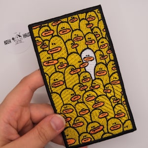 May include: A rectangular embroidered patch featuring a dense pattern of yellow cartoon ducks with orange beaks, with one single white duck positioned near the center. The patch has a thick black border and a textured, stitched surface throughout.