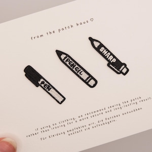 Peut inclure: Trois écussons brodés en noir et blanc en forme de stylo, de crayon et de marqueur, chacun portant son nom. Les écussons sont présentés sur une carte blanche avec le texte from the patch haus et des instructions d'application.