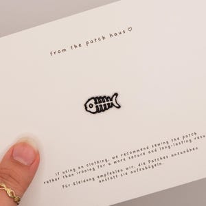 Peut inclure: Un petit écusson brodé noir en forme d'arête de poisson centré sur une carte blanche. La carte porte le texte from the patch haus avec un symbole de cœur en haut et des instructions sur la couture par rapport au repassage en bas.