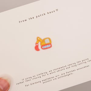 May include: A small embroidered patch shaped like a yellow construction excavator with a red bucket and blue tracks. The patch is centered on a white card with the text from the patch haus and instructions for sewing or ironing the patch.