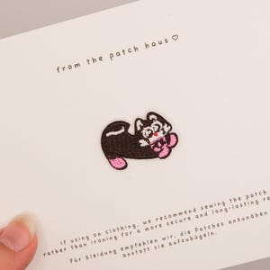 May include: An embroidered patch featuring a black and white cat with pink accents, displayed on a white card with the text from the patch haus and instructions recommending sewing the patch onto clothing for a secure and long-lasting result.