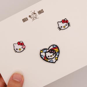 Puede incluir: Tres parches bordados con el personaje Hello Kitty, incluyendo dos pequeños retratos de cabeza con lazos rojos y un parche en forma de corazón con un arcoíris y nubes. El texto THE PATCH HAUS está impreso sobre los parches en la tarjeta blanca.