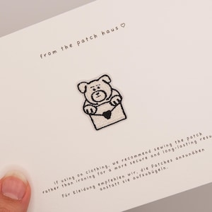 以下が含まれることがあります： 白いカードの中央に配置された、ハート付きの封筒を持つクマの刺繍ワッペン。カードには「from the patch haus」という文字と、衣類にしっかりと取り付けるために縫い付けることを推奨する説明書きが記載されています。