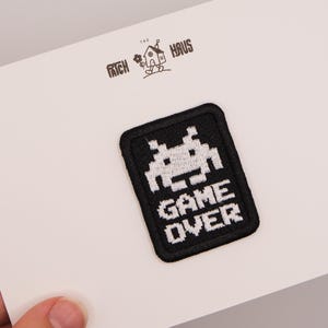 May include: A rectangular black embroidered patch featuring a white pixelated alien graphic above the text GAME OVER. The patch is displayed on a white card with the logo THE PATCH HAUS printed in black at the top center of the card.