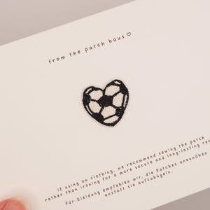 Puede incluir: Un pequeño parche bordado en forma de corazón con un patrón de balón de fútbol en blanco y negro. El parche está centrado en una tarjeta blanca con el texto from the patch haus e instrucciones para coser o planchar el parche en la ropa.