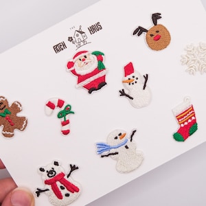 May include: A collection of embroidered Christmas-themed patches on a white card, including a gingerbread man, candy cane, Santa Claus, reindeer, snowman, snowflake, stocking, and a polar bear wearing a red scarf, with the text THE PATCH HAUS.