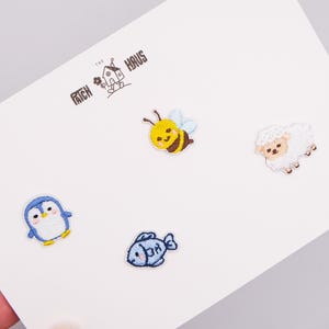 May include: A set of four small embroidered patches on a white card featuring the text THE PATCH HAUS. The designs include a yellow and black bee, a white fluffy sheep, a blue and white penguin, and a blue fish, all with stitched outlines.