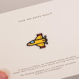 Puede incluir: Un parche bordado con forma de avión de combate amarillo con cabina azul claro y aletas traseras gris oscuro. El parche está centrado en una tarjeta blanca con el texto from the patch haus e instrucciones para coser el parche en la ropa.
