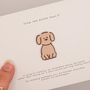 May include: A light brown embroidered dog patch on a white card. The card features the text from the patch haus with a heart symbol, and instructions in English and German recommending sewing the patch onto clothing for a secure result.