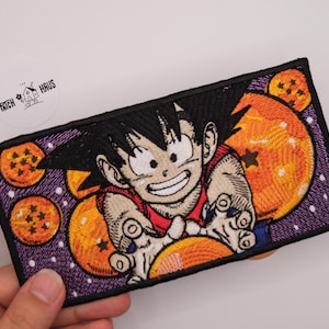 Puede incluir: Un parche rectangular bordado que muestra un personaje de dibujos animados con cabello negro puntiagudo y camisa roja, rodeado de cuatro esferas naranjas con estrellas rojas sobre un fondo morado con puntos blancos, con un borde negro.