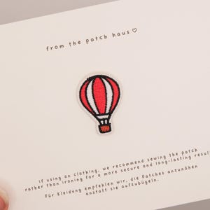 May include: An embroidered hot air balloon patch featuring red and white vertical stripes with a black outline and basket. The patch is centered on a white card with the text from the patch haus and instructions for sewing the patch onto clothing.