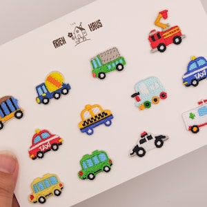 May include: A collection of twelve small embroidered vehicle patches arranged on a white card labeled THE PATCH HAUS. The set includes a red fire truck, blue taxi, green truck, yellow cement mixer, ambulance, and various other colorful cars and trucks.