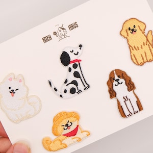 May include: Five embroidered dog patches on a white card with the text PATCH HAUS. The designs include a white fluffy dog, a black and white spotted dog, a golden dog, a brown and white spaniel, and a tan dog wearing a hat and red scarf.
