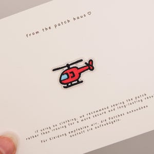 Könnte beinhalten: Ein kleiner roter Hubschrauber-Aufnäher mit hellblauem Fenster und schwarzen Details, zentriert auf einer weißen Karte. Die Karte enthält den Text "from the patch haus" sowie Anweisungen, den Aufnäher für ein sicheres Ergebnis anzunähen.