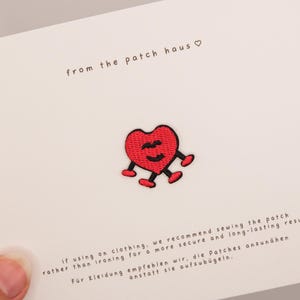 Puede incluir: Un parche bordado en forma de corazón rojo con rasgos faciales y extremidades cosidas en negro, centrado en una tarjeta blanca. La tarjeta incluye el texto from the patch haus e instrucciones en inglés y alemán sobre cómo coser el parche.
