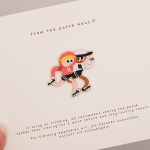 Puede incluir: Un parche bordado que muestra a dos figuras de dibujos animados patinando, una con una chaqueta rosa con capucha con una R blanca y la otra con un conjunto blanco y negro. El parche se presenta sobre una tarjeta blanca con texto e instrucciones.