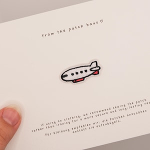 Könnte beinhalten: Ein kleiner gestickter Aufnäher in Form eines weißen Flugzeugs mit schwarzen Umrissen und roten Akzenten, zentriert auf einer weißen Karte. Die Karte enthält den Text from the patch haus mit einem Herzsymbol und Anweisungen zum Annähen des Patches.