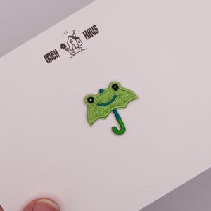 Könnte beinhalten: Ein gestickter Aufnäher in Form eines hellgrünen Frosches, der wie ein Regenschirm gestaltet ist, mit einem dunkelgrünen Griff und schwarzen Augen. Der Aufnäher ist auf einer weißen Karte mit dem Text THE PATCH HAUS und einer Hausgrafik.