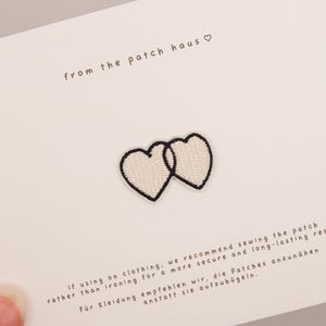 Puede incluir: Un parche bordado que muestra dos corazones entrelazados con hilo de color crema y un borde negro. El parche está sujeto a una tarjeta blanca impresa con el texto from the patch haus e instrucciones para coser el parche en la ropa.