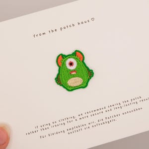 Könnte beinhalten: Ein gestickter Aufnäher in Form eines grünen, einäugigen Monsters mit orangen Ohren und beigem Bauch. Der Aufnäher ist auf einer weißen Karte befestigt, die den Text from the patch haus sowie Anweisungen zum Annähen auf Englisch und Deutsch enthält.