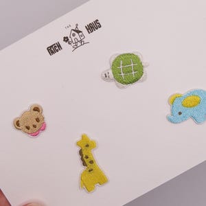 以下が含まれることがあります： THE PATCH HAUSの文字が記された白い台紙に、4つの刺繍ワッペンが貼られています。ワッペンは、緑色のカメ、黄色い耳をした水色のゾウ、茶色の斑点がある黄色のキリン、ピンクの襟をしたベージュのクマの形をしています。