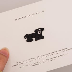 May include: A black embroidered dog patch centered on a white card. The card features the text from the patch haus with a heart symbol, and instructions in English and German regarding sewing the patch onto clothing for a secure result.