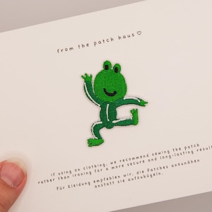 Peut inclure: Un écusson brodé représentant une grenouille verte dans une pose dynamique. L'écusson est fixé sur une carte blanche avec le texte from the patch haus et des instructions en anglais et en allemand sur la couture de l'écusson sur un vêtement.