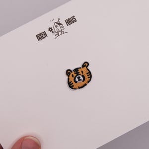 Pode incluir: Um emblema bordado apresentando uma cabeça de tigre de desenho animado com detalhes em fio laranja e preto. O emblema é exibido em um cartão branco impresso com o texto THE PATCH HAUS e um pequeno desenho de linha de uma casa com uma flor.