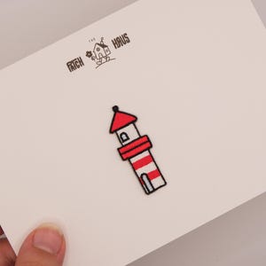 Könnte beinhalten: Ein gestickter Leuchtturm-Aufnäher mit roten und weißen horizontalen Streifen, einem roten Dach und einem kleinen Fenster. Der Aufnäher ist auf einer weißen Karte befestigt, die mit dem Text THE PATCH HAUS und einer Strichzeichnung eines Hauses bedruckt ist.