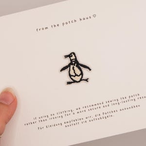 Op de afbeelding: Een kleine geborduurde pinguïn patch in zwart en wit garen, gecentreerd op een witte kaart. De kaart bevat de tekst from the patch haus en instructies in het Engels en Duits over het naaien van de patch op kleding voor een duurzaam resultaat.